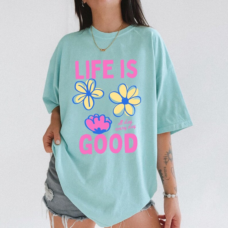 May include: A pale blue t-shirt featuring the pink text "LIFE IS GOOD" and three flower illustrations. The shirt has a crew neck and short sleeves. The words "all day everyday" are also printed on the shirt.