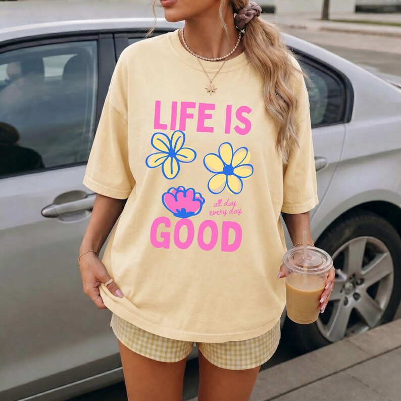 May include: A pale yellow t-shirt with the words "LIFE IS GOOD" in pink and blue, and flower graphics. The shirt is worn with plaid shorts. A person is holding a drink in a clear cup.