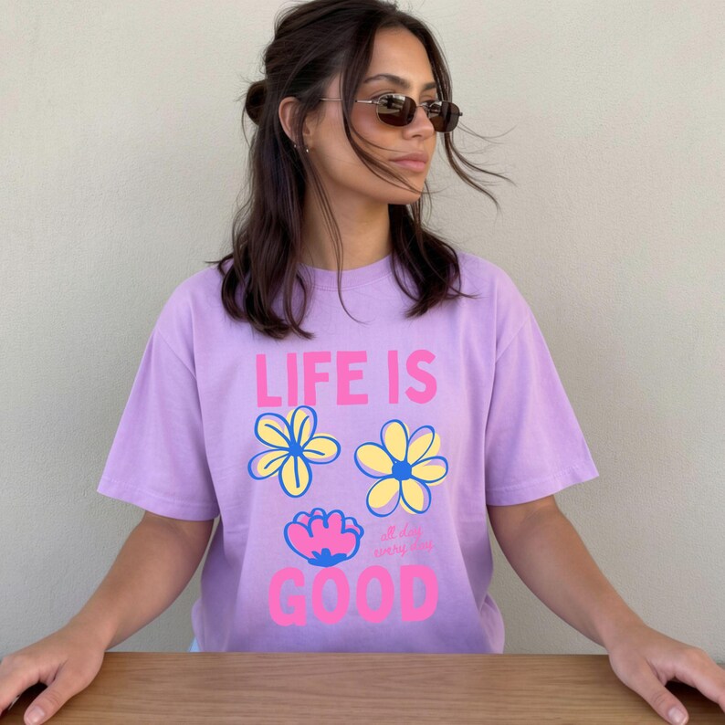 May include: A lilac t-shirt featuring the text "LIFE IS GOOD" in pink and white, and three flower illustrations in yellow, blue, and pink. The shirt has short sleeves and a relaxed fit.