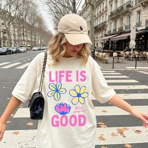 May include: A cream-coloured t-shirt with the text "LIFE IS GOOD" in pink and blue, and flower graphics. The outfit includes a beige baseball cap with a navy blue logo. A black purse with a chain strap is also visible.