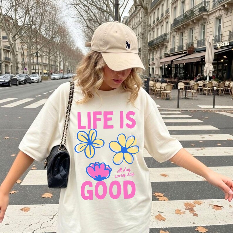May include: A cream-coloured t-shirt with the text "LIFE IS GOOD" in pink and blue, and flower graphics. The outfit includes a beige baseball cap with a navy blue logo. A black purse with a chain strap is also visible.