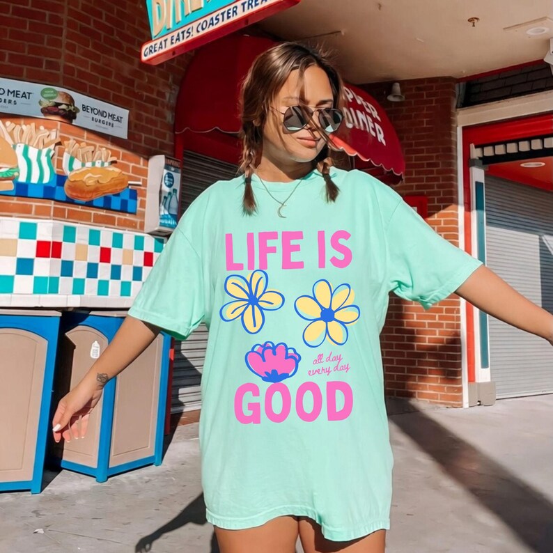 May include: A pale green t-shirt with the words "LIFE IS GOOD" in pink and white, and flower graphics. The shirt also has the words "all day every day" in a smaller font. The shirt is worn by a person wearing sunglasses.