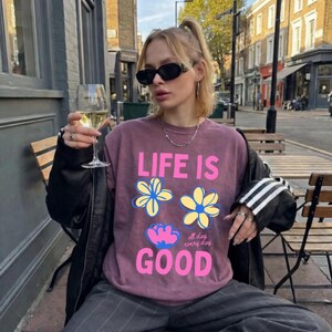May include: A mauve sweatshirt with the text "LIFE IS GOOD" and floral designs. The person is wearing sunglasses and holding a glass of white wine. The sweatshirt is paired with grey trousers and a black jacket.