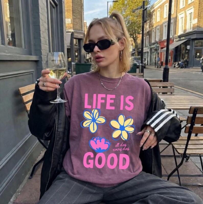 May include: A mauve sweatshirt with the text "LIFE IS GOOD" and floral designs. The person is wearing sunglasses and holding a glass of white wine. The sweatshirt is paired with grey trousers and a black jacket.
