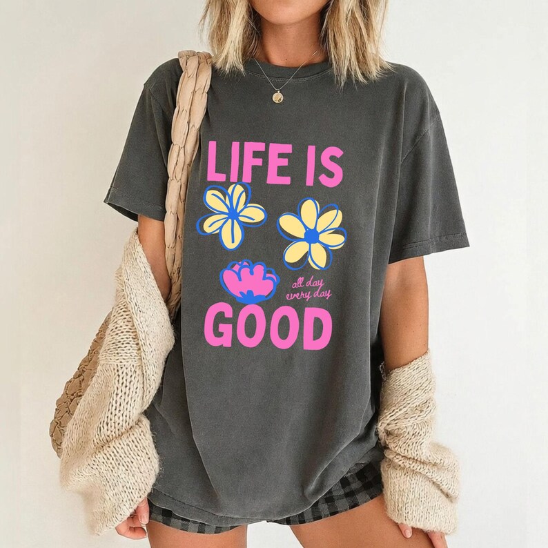 May include: Dark grey t-shirt featuring the words "LIFE IS GOOD" in pink, with blue and yellow flower graphics. The phrase "all day every day" is below the flowers. The shirt has a relaxed, oversized fit.