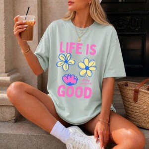 May include: A pale green t-shirt featuring the text "LIFE IS GOOD" in pink, with blue and yellow flower illustrations. The shirt also includes the words "all day every day". The t-shirt has a relaxed, oversized fit.