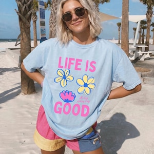 May include: A pale blue t-shirt with the words "LIFE IS GOOD" in pink, and flower designs. The person is wearing sunglasses and colourful shorts. The background features palm trees and a beach.