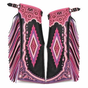 Puede incluir: Un par de chaparreras de cuero negro con grabados florales en rosa y morado, un patrón geométrico central en forma de diamante y largos flecos en los laterales. Cada pierna incluye una hebilla superior y el logo NEXT STEP GEAR.