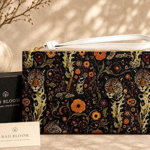 Può includere: Una pochette rettangolare con cinturino da polso, caratterizzata da uno sfondo nero con un motivo ripetuto di leopardi, fiori arancioni e fogliame verde. La pochette presenta una cerniera dorata e un cinturino bianco. Il testo BAD BLOOM è visibile.