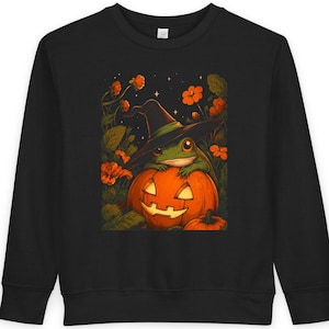 Puede incluir: Una sudadera negra de manga larga con un estampado gráfico de una rana verde con un sombrero de bruja negro, descansando sobre una calabaza naranja tallada rodeada de flores naranjas y hojas verdes bajo un cielo nocturno estrellado.
