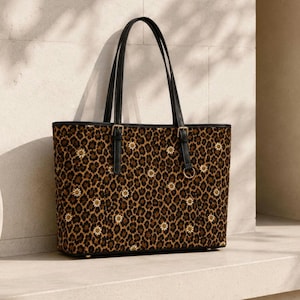 Può includere: Una borsa tote strutturata con motivo leopardato marrone e nero, arricchita da piccoli fiori di margherita bianchi. La borsa presenta due sottili tracolle nere con fibbie dorate e un dettaglio ad anello dorato sulla parte anteriore.