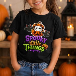 Può includere: Una t-shirt nera a maniche corte con la grafica di un fantasma bianco dentro una zucca arancione, affiancato da due pipistrelli neri. Sotto la grafica, il testo "SPOOKY LITTLE THINGS" in viola e verde, con una piccola icona di zucca.