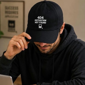 Può includere: Un cappellino da baseball nero con testo ricamato in bianco che recita 404 MOTIVATION NOT FOUND, accompagnato da una piccola grafica di un monitor per computer e un'icona del cursore centrata sul pannello frontale.