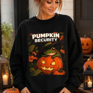 Può includere: Una felpa nera a maniche lunghe con una stampa grafica di una rana verde che indossa un cappello da strega nero, seduta su una zucca arancione intagliata. Il testo PUMPKIN SECURITY è stampato in bianco sopra la rana tra fiori e foglie.
