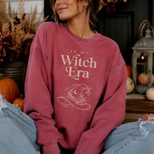 Può includere: Una felpa girocollo color rosa antico con il testo "IN MY WITCH ERA" in un carattere serif color crema, accompagnata da un'illustrazione a linee di un cappello da strega decorato con stelle celesti e motivi floreali vorticosi.
