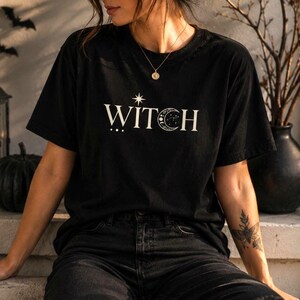 Può includere: Una t-shirt nera a maniche corte con la parola WITCH stampata in bianco sul petto. La lettera I è sormontata da una stella a quattro punte e la lettera C è sostituita da una luna crescente con piccoli motivi di stelle celesti all'interno.