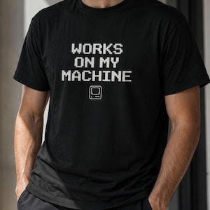 Può includere: Una t-shirt nera a maniche corte con il testo pixelato bianco "WORKS ON MY MACHINE" centrato sul petto, posizionato sopra una piccola icona a linee bianche di un monitor per computer vintage.