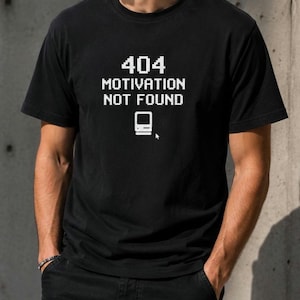 Può includere: Una t-shirt nera a maniche corte con testo bianco pixelato che recita 404 MOTIVATION NOT FOUND, accompagnata da una piccola grafica di un monitor per computer vintage e un'icona del cursore del mouse centrata sul petto del capo.