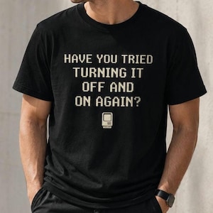 Può includere: Una t-shirt nera a maniche corte con il testo pixelato bianco "HAVE YOU TRIED TURNING IT OFF AND ON AGAIN?" centrato sul petto, posizionato sopra una piccola e semplice illustrazione grafica bianca di un monitor per computer vintage.