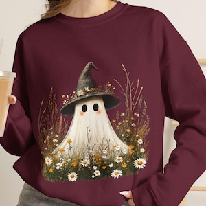 Può includere: Una felpa girocollo bordeaux con una stampa grafica raffigurante un fantasma bianco che indossa un cappello a punta nero decorato con piccoli fiori gialli e bianchi. Il fantasma è circondato da un campo di erba alta e margherite bianche.
