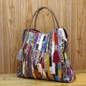 Op de afbeelding: Een veelkleurige handtas met een verticaal patchworkontwerp met diverse texturen, waaronder dierenprints en metallic afwerkingen. De tas heeft twee gevlochten handvatten, een lichtgrijze kwast aan de linkerkant en een donkerrode kwast rechts.