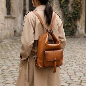 May include: A brown leather backpack with a front pocket and adjustable straps. The backpack is worn over the shoulders. The person is wearing a tan trench coat. The background is a cobblestone street.