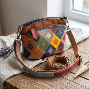 May include: A multi-colored leather crossbody bag featuring a geometric diamond patchwork pattern in shades of yellow, green, blue, and orange. The bag includes a zippered top, a red leather zipper pull, and an adjustable canvas shoulder strap.