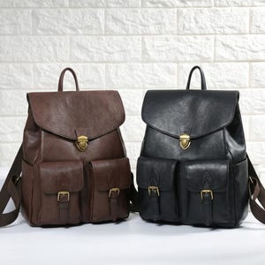 May include: Two leather backpacks, one brown and one black, are displayed side-by-side. Each backpack features a flap closure, two front pockets with buckles, and a top handle. The brown backpack has brown straps, while the black backpack has black straps.