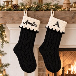 Op de afbeelding: Twee zwarte kerstsokken van kabelgebreid materiaal met witte geribbelde boorden en een zwarte schulprand. De linker sok is geborduurd met de naam Amelia in zwart cursief schrift en de rechter sok toont de letter A in een zwart schreeflettertype.