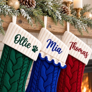 Könnte beinhalten: Drei gestrickte Weihnachtsstrümpfe mit Zopfmuster in Grün, Blau und Rot. Jeder Strumpf hat ein weißes Rippbündchen, das mit Namen in Schreibschrift personalisiert ist: Ollie mit einer grünen Pfote, Mia in blauem Text und Thomas in rotem Text.