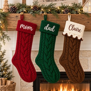 Könnte beinhalten: Drei gestrickte Weihnachtssocken mit Zopfmuster hängen in einer Reihe. Die erste ist rot mit weißem Text mom, die zweite grün mit weißem Text dad und die dritte braun mit weißem Bündchen mit Wellenrand und braunem Text Claire.