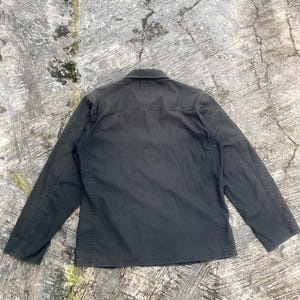 May include: A dark charcoal gray long-sleeve button-up shirt featuring a standard collar, a horizontal back yoke, and visible vertical seam detailing along the sides and sleeves. The garment is displayed flat, showcasing its solid color and construction.