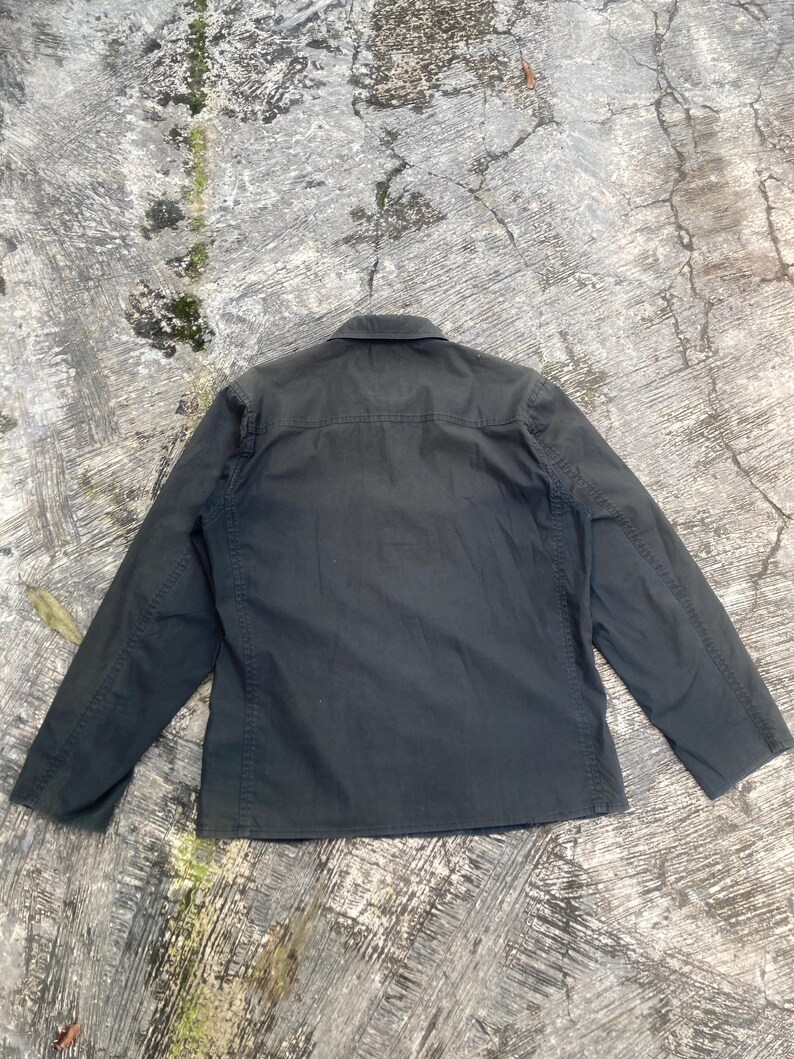 May include: A dark charcoal gray long-sleeve button-up shirt featuring a standard collar, a horizontal back yoke, and visible vertical seam detailing along the sides and sleeves. The garment is displayed flat, showcasing its solid color and construction.