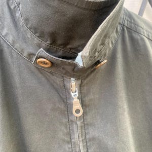 May include: A dark olive green jacket featuring a high stand-up collar with a button closure tab. The front includes a central metal zipper with a YKK pull tab. The fabric has a matte finish and visible stitching along the collar and placket.