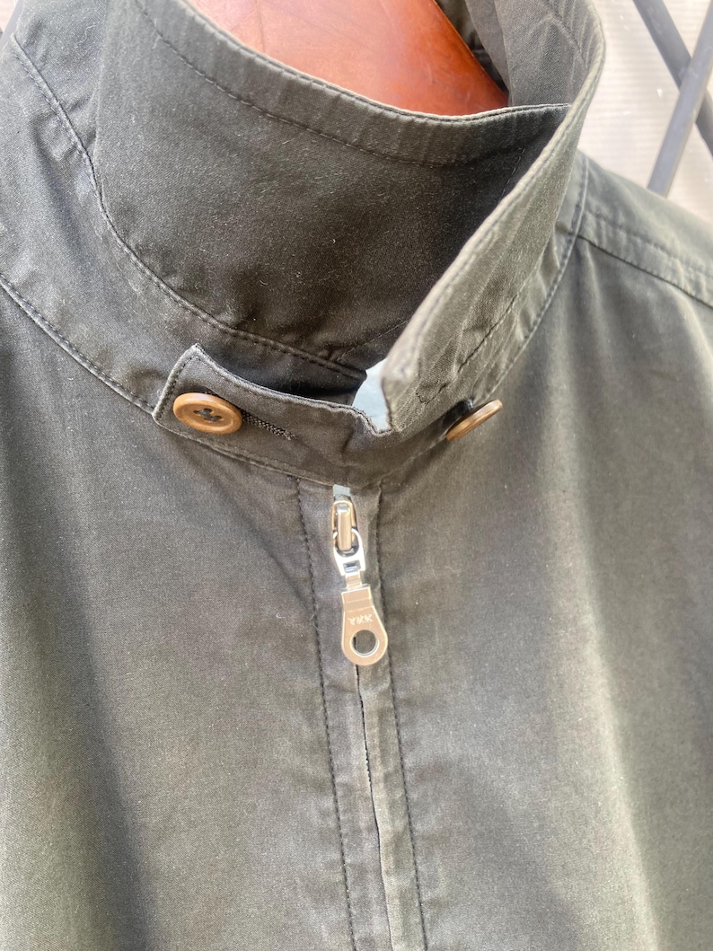 May include: A dark olive green jacket featuring a high stand-up collar with a button closure tab. The front includes a central metal zipper with a YKK pull tab. The fabric has a matte finish and visible stitching along the collar and placket.