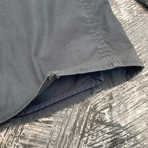 May include: A dark gray jacket sleeve featuring a visible frayed area along the bottom hem edge. The garment is constructed with reinforced stitching along the seams and a smooth fabric texture, showing signs of wear at the cuff opening.