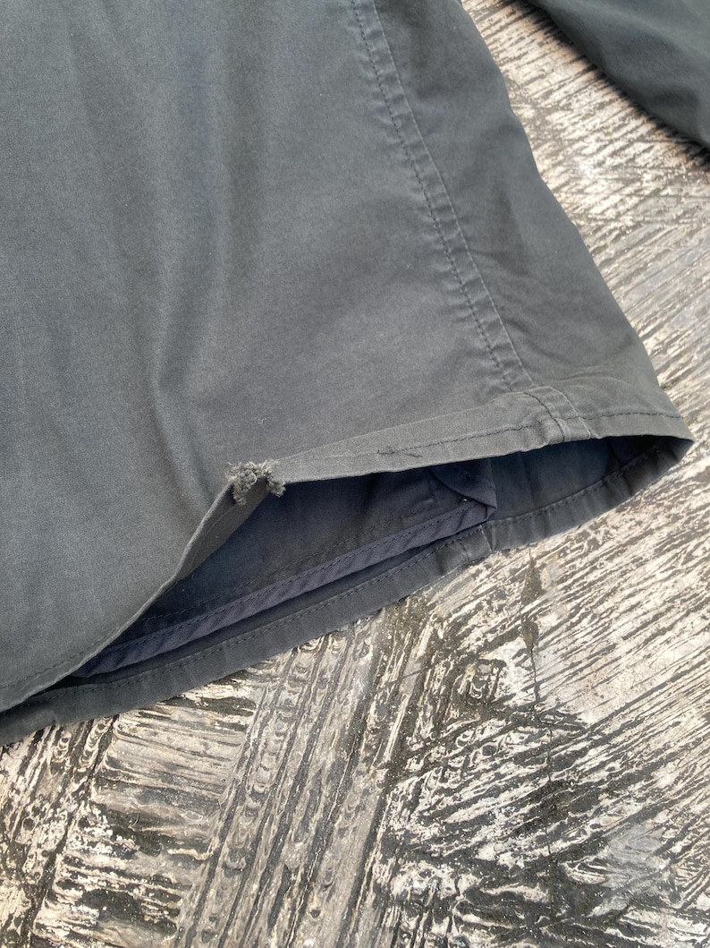 May include: A dark gray jacket sleeve featuring a visible frayed area along the bottom hem edge. The garment is constructed with reinforced stitching along the seams and a smooth fabric texture, showing signs of wear at the cuff opening.