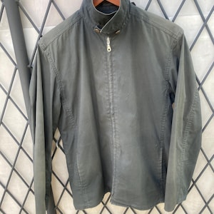 May include: A dark olive green jacket featuring a high collar with a snap button closure, a full-length front zipper with a metallic pull, long sleeves, and two side pockets. The jacket is displayed on a wooden hanger against a metal grid.