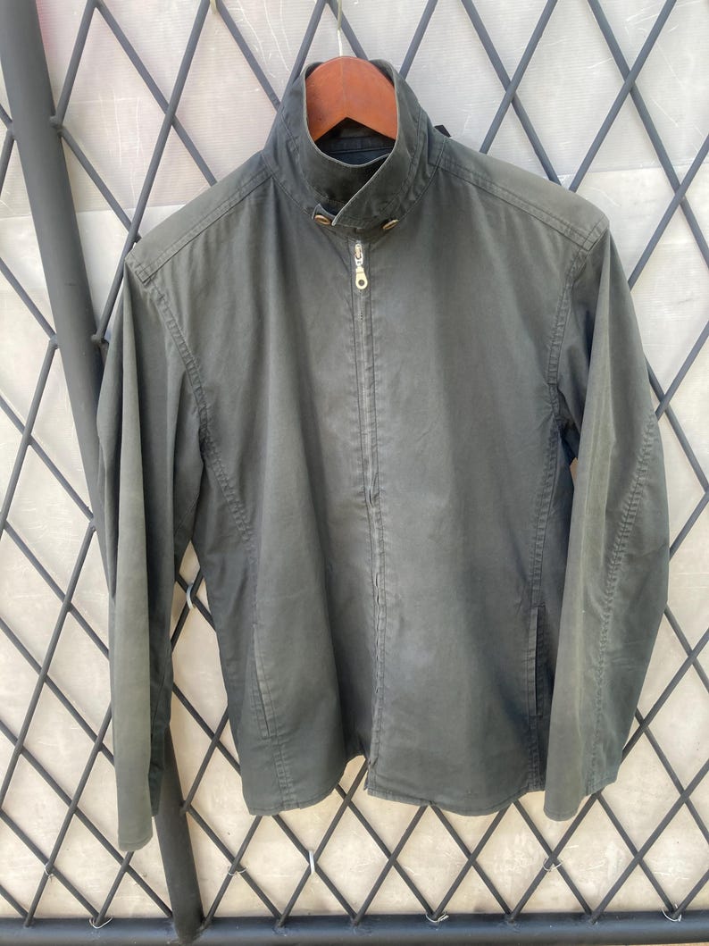 May include: A dark olive green jacket featuring a high collar with a snap button closure, a full-length front zipper with a metallic pull, long sleeves, and two side pockets. The jacket is displayed on a wooden hanger against a metal grid.