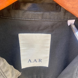 May include: A dark olive green shirt featuring a white rectangular interior neck label with the text A.A.R printed in dark blue. The garment includes a matching dark olive green fabric loop at the collar and a small white size tag on the right side.