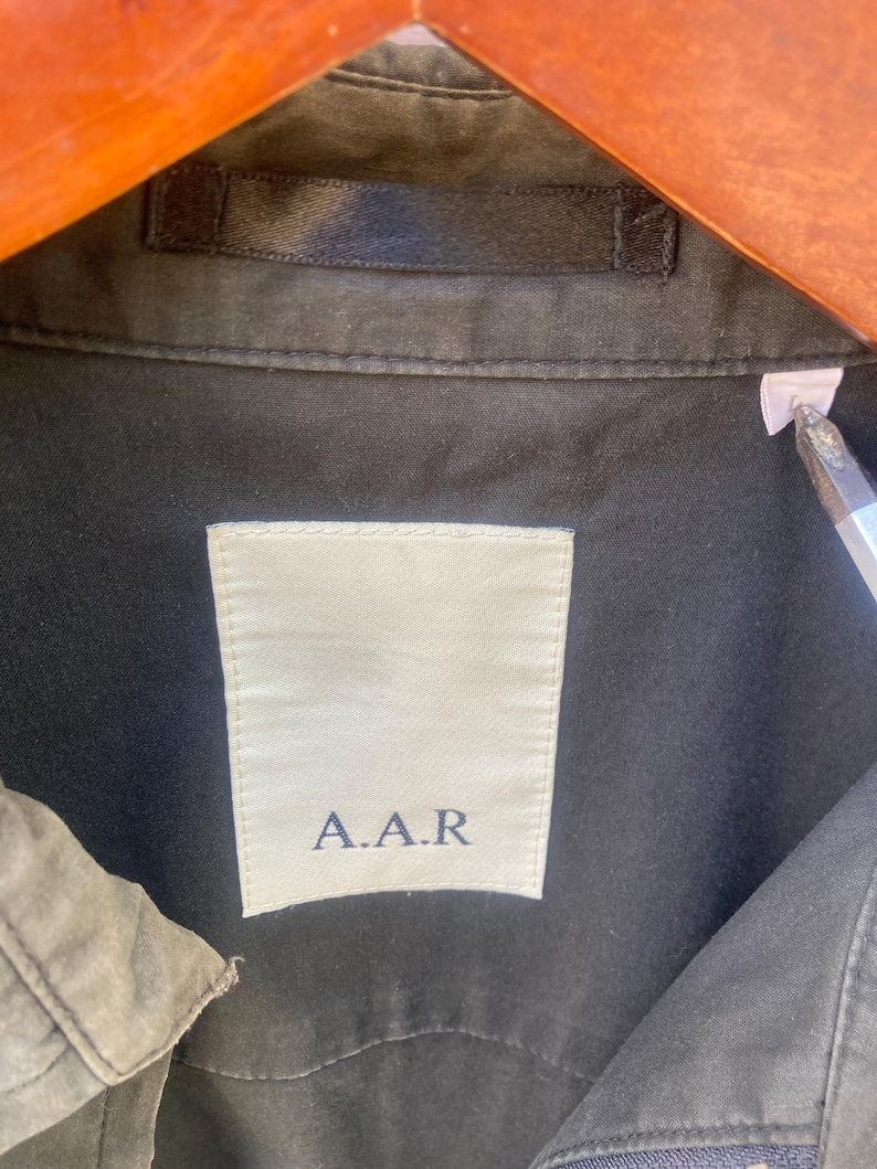 May include: A dark olive green shirt featuring a white rectangular interior neck label with the text A.A.R printed in dark blue. The garment includes a matching dark olive green fabric loop at the collar and a small white size tag on the right side.