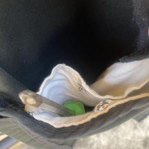 May include: A black fabric pocket with a white interior lining containing a green-handled tool with a metallic hexagonal shaft. The pocket features a small silver metal snap button fastener near the top edge of the white fabric lining.