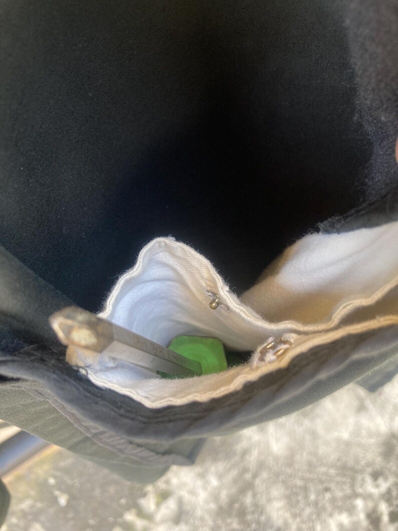 May include: A black fabric pocket with a white interior lining containing a green-handled tool with a metallic hexagonal shaft. The pocket features a small silver metal snap button fastener near the top edge of the white fabric lining.