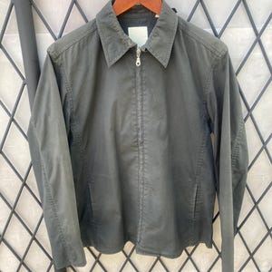 May include: A dark olive green zip-up jacket featuring a classic pointed collar, long sleeves, and two side pockets. The garment has a central silver-toned metal zipper closure and a plain, unbranded white label visible at the inner neckline.