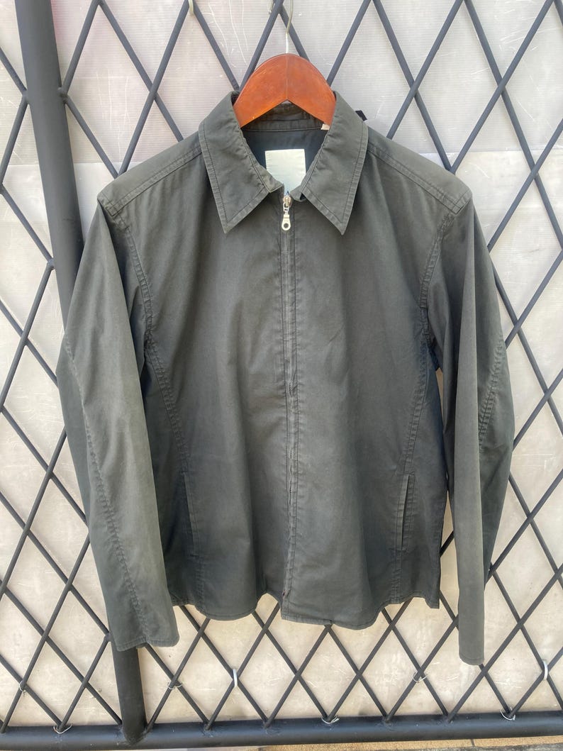 May include: A dark olive green zip-up jacket featuring a classic pointed collar, long sleeves, and two side pockets. The garment has a central silver-toned metal zipper closure and a plain, unbranded white label visible at the inner neckline.