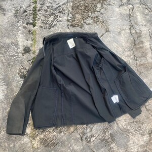 May include: A dark charcoal gray jacket with a full-length front zipper, long sleeves, and two large front patch pockets. The interior features a white brand label with the text AAK at the collar and a white care tag on the inner side seam.