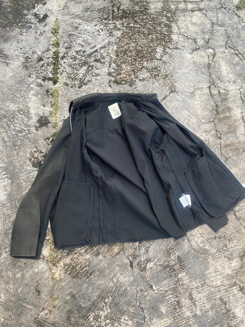 May include: A dark charcoal gray jacket with a full-length front zipper, long sleeves, and two large front patch pockets. The interior features a white brand label with the text AAK at the collar and a white care tag on the inner side seam.