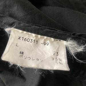 May include: A black garment featuring a white fabric care label with the text X160311 -99, L, and Japanese characters indicating 95% cotton and 5% polyurethane. The label is stitched onto the dark fabric with visible frayed white threads at the edges.