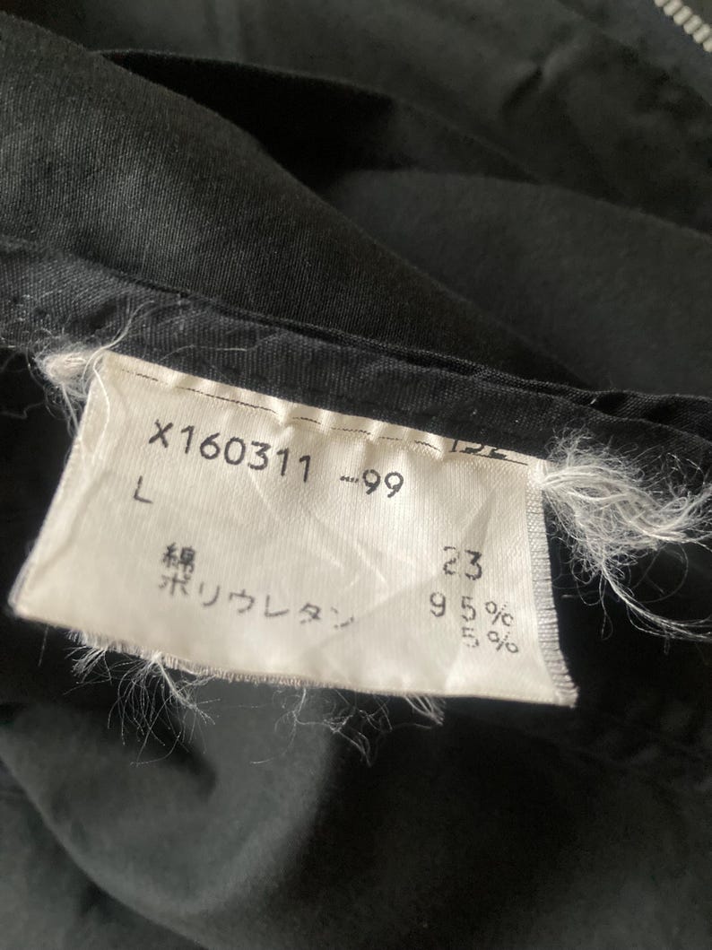 May include: A black garment featuring a white fabric care label with the text X160311 -99, L, and Japanese characters indicating 95% cotton and 5% polyurethane. The label is stitched onto the dark fabric with visible frayed white threads at the edges.