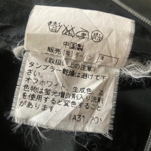 May include: A white rectangular garment care label sewn onto black fabric. The label features black Japanese text, including instructions to avoid tumble drying and warnings about color changes with fluorescent detergents, along with laundry symbols.